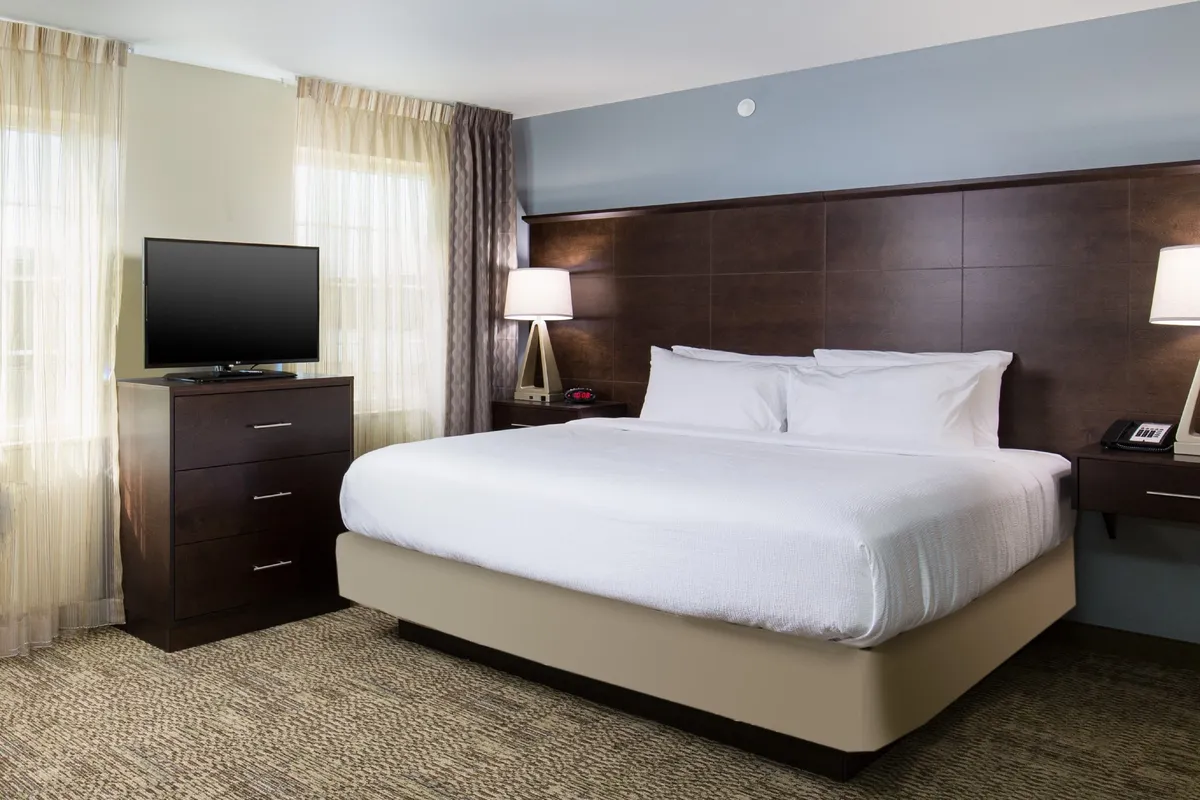 A peaceful bedroom featuring a plush King-sized bed, a wall-mounted TV, a dresser, and elegant window treatments. The soft lighting and calming decor create a serene atmosphere ideal for restful nights.