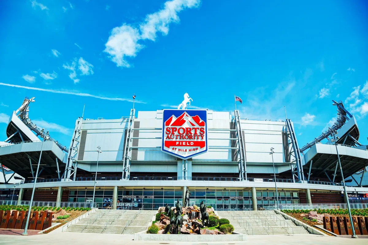 A view of the impressive Sports Authority Field, showcasing the stadium’s architecture, statues in the foreground, and signage. The scene conveys excitement and the spirit of large-scale events and gatherings.