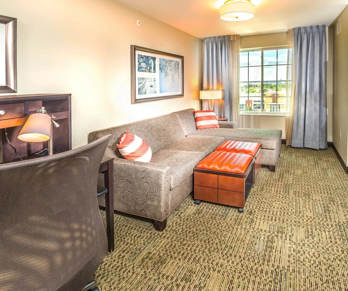 The image showcases a spacious, sunlit suite interior with a plush sofa positioned by a large window offering skyline views, tasteful modern artwork, and a dedicated desk area—ideal for both relaxation and productivity. The setting encourages guests to unwind, enjoy city views, or catch up on work in a comfortable, stylish environment.