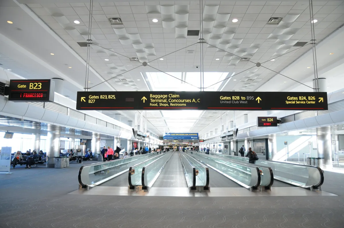 The image shows a bustling airport interior featuring a moving walkway, clear signage for gates, and travelers waiting or moving through the space. The atmosphere is busy yet organized, with accessible pathways and multiple points of entry visible. The scene highlights the anticipation of travel and the movement of people, including travelers with varying needs.
