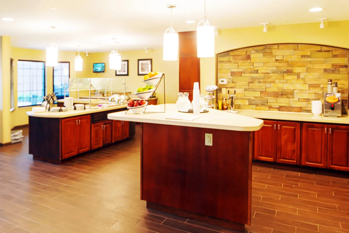 A welcoming dining area anchored by a central island with a fruit display, stone accent wall, and hanging light fixtures. Guests are shown enjoying breakfast, coffee, or relaxing with a book, capturing the lively yet relaxed atmosphere of mornings or communal meals.