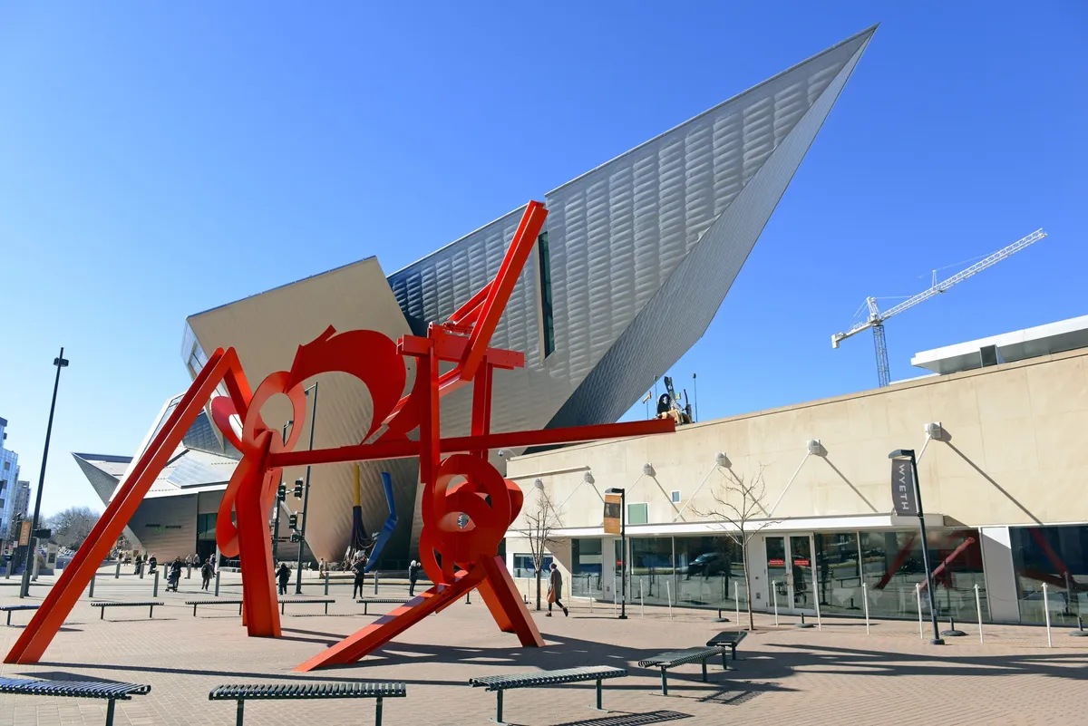 An outdoor scene featuring a bold red sculpture and modern architecture, with people walking and gathering in the area. The setting feels lively and welcoming, suggesting a vibrant cultural or shopping district.