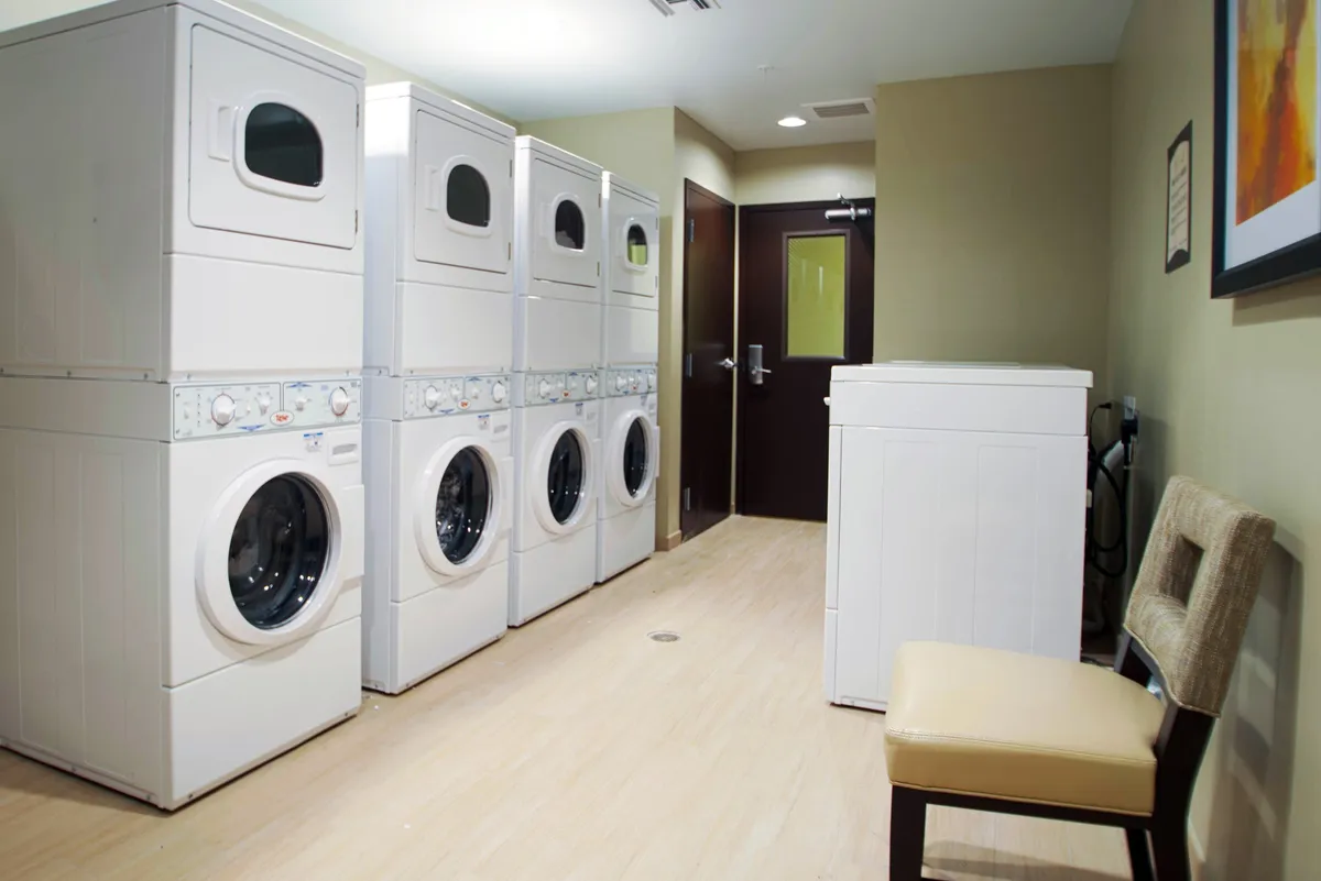 A practical laundry room with stacked washer-dryer units and a single washer, paired with a comfortable chair for waiting. The setting is clean, functional, and welcoming, with guests completing laundry and occasionally chatting with neighbors.
