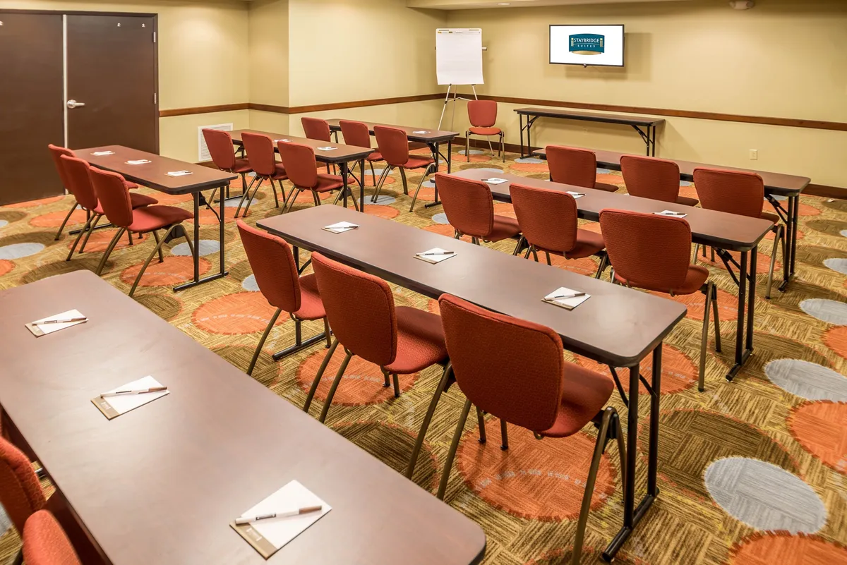 A spacious meeting room with rows of tables and vibrant red chairs facing a large presentation screen, set up for collaborative work, workshops, or group meetings. The orderly arrangement fosters a focused, professional environment.